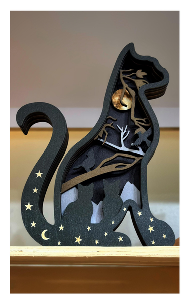 A piece of handcrafted art displayed on a shelf in a gift shop. It’s a thick, flat board cut into the shape of a cat sitting in profile, painted black, with a crescent moon and stars scattered along its paws and tail. The interior of the profile is also cut out, at a more shallow depth, retaining a backing board. This creates space for a diorama of cat and bird silhouettes, branches, distant mountains, and a golden full moon, all made out of flat cutouts positioned at different depths. The artwork perhaps suggests the interior life of a cat, dreaming of birds and wide spaces and moonlight.