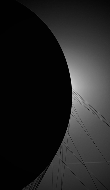 A silhouette of a large circular object, possibly a balloon or dome, against a gradient background. Thin lines radiate from the object, suggesting strings or cables. The image is predominantly in black and white, creating a dramatic contrast.