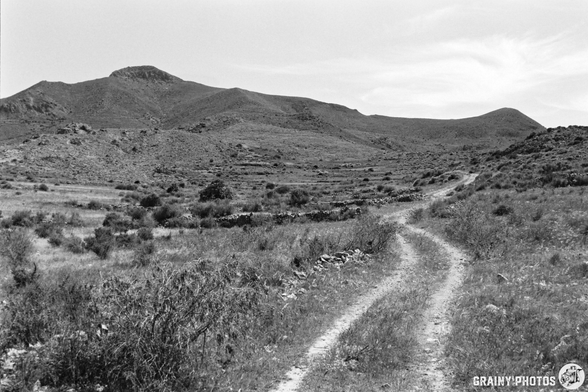 A winding dirt path leads through a grassy landscape, with rolling hills and sparse vegetation. The scene is captured in black and white, highlighting the textures of the terrain and the contrast between light and shadow.