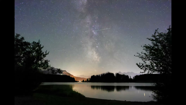 A 6 second time lapse of a view across a large lake with the night sky with the Milky Way and countless airplanes moving by. The scene is framed by the silhouettes of some bushes. The orange glow of the town of Füssen near the horizon. Faint mountain ridges further back.