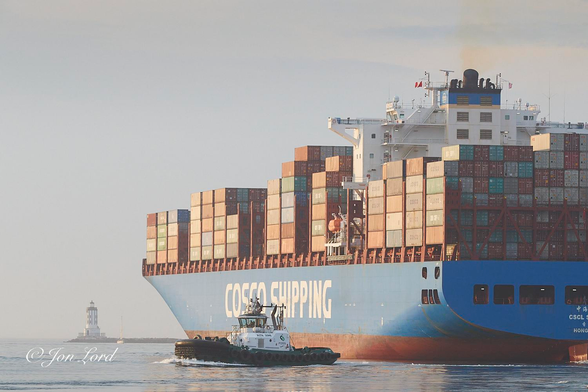 This is a colour photo in landscape format of a large container ship underway and leaving port at sunrise with a tug departing to one side and a lighthouse ahead. Port of Los Angeles, California (2018).

Extending upwards from the base of the photo to less than the quarter up level is a calm and mirror like, light blue, harbour water. Stretching from the right edge of the photo, going left and through the centre to towards the right side of the photo is our container ship. The ship is viewed obliquely with the stern and port (left) side in view and is underway from right to left. The ship fills three-quarters of the height of the photo, three-quarters of the width and is viewed from about ten metres away. Only the port side of the stern is in view, the starboard side is beyond the right edge of the image. The vessel is riding high in the water with about two metres of red antifouling visible above the waterline. Above the hull is a medium blue with the words 'COSCO SHIPPING' in large white lettering, the shipping line's name (China Overseas Shipping Company). On the stern, and partially out of view, is the vessel's name in both Chinese characters and English: 'CSCL AUTUMN' and the port of registry: 'Hong Kong'. Above the decks are thousands of multicoloured 'forty foot' shipping containers. The still low morning sun illuminating the containers and hull. The containers are stacked eighteen across and seven high in places with randomly spaced gaps. About a quarter of the way forward from the stern is the white superstructure containing the accommodation, wheelhouse and funnel on top. On the short blue, square funnel are numerous black exhaust pipes with one large, perhaps two metres in diameter, emitting a wisp of brown to orange smoke from her main diesel engine.  While the ship is underway at only a few knots there is hardly any wake behind. To the port side and abeam the superstructure is a large tug angled at right angles to the ship, only a few metres away and underway to the left. In the lower left corner and ahead of the bow of the ship is a white cylindrical lighthouse with vertical black stripes extending upwards to the lantern. Stretching to the right of the lighthouse is the harbour breakwater with a calm Pacific Ocean beyond. In the background is an ill defined horizon with a hazy sky above and beyond. 