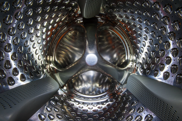 Bildbeschreibung (Deutsch) Das Bild zeigt einen tiefen Blick in das Innere einer glänzenden Waschtrommel aus Edelstahl. Die Wände sind mit einem komplexen Muster aus kleinen, diamantförmigen Vertiefungen und Abflusslöchern bedeckt, die das Licht reflektieren. Drei perforierte, graue Mitnehmer-Paddel ragen von den Seiten in die Trommel. Der hintere Teil der Trommel hat ein sternförmiges Design, das eine optische Tiefe erzeugt. Image Description (English) The image displays a deep perspective looking into the interior of a gleaming stainless steel washing machine drum. The walls are covered in a complex pattern of small, diamond-shaped dimples and drain holes that reflect light. Three perforated gray agitator paddles protrude from the sides into the drum. The rear part of the drum has a star-shaped design, creating visual depth. 
