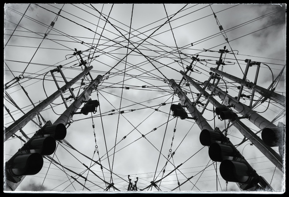 A monochrome picture looking up at a cloudy sky from the corners of an intersection with many overhead power, electric bus, and communication wires. Six power poles shoot up from the lower corners of the picture. Dozens of criss-crossing wires fill the frame.

This is a merge of three separate photos taken while wandering around the corners of a busy intersection on the edge of the Mission. The merge is done in Photoshop using the Dark layer blend. Dark Mode allows darker pixels from upper layers to overlay lighter pixels on lower layers. 

This Dark Mode technique to merge pictures doesn't usually work as well as you would expect it if a scene has depth, as it can cause dark areas in the distance to seemingly jump forward because they overlay lighter, closer objects. In this case it didn't matter... probably because the poles and wires in the foreground were all about the same amount of darknese while the brighter pixels, which got overlaid, were all from distant clouds. In other words, the bright gray sky remained where it should: in the distance.

And that explains why I had to wait for an overcast day to make this work.